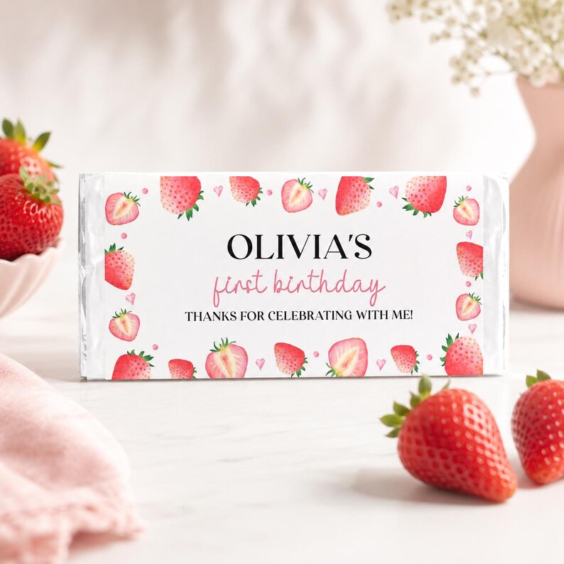 May include: A rectangular candy bar wrapper with a white background featuring a pattern of red strawberries and small pink hearts. The text reads OLIVIA'S first birthday THANKS FOR CELEBRATING WITH ME! in black and pink serif and script fonts.