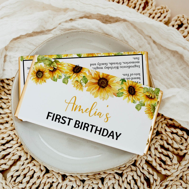 May include: A rectangular chocolate bar wrapper featuring a watercolor sunflower border at the top. The text reads Amelia's FIRST BIRTHDAY in gold and black lettering. The back of the wrapper lists ingredients including birthday magic, memories, friends, family, and lots of fun.