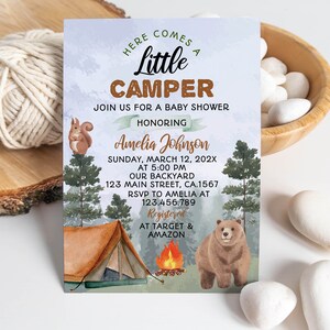 Op de afbeelding: Een rechthoekige uitnodigingskaart voor een babyshower met een bosthema, inclusief een bruine beer, een eekhoorn, een tent en een kampvuur. De tekst luidt: Here Comes A Little Camper, Join Us For A Baby Shower Honoring Amelia Johnson, March 12, 202X.