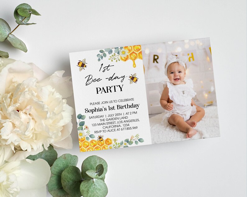 May include: A bee-themed first birthday party invitation with a photo of a baby girl wearing a white dress. The invitation features a yellow honeycomb, bees, and green leaves. The text on the invitation reads "1st Bee-day Party" and "Sophia's 1st Birthday".