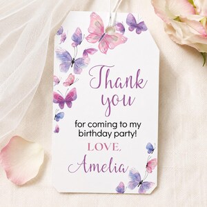 May include: A rectangular white gift tag with a hole at the top, decorated with watercolor-style purple and pink butterflies. The text reads Thank you for coming to my birthday party! LOVE, Amelia in purple and black script and sans-serif fonts.