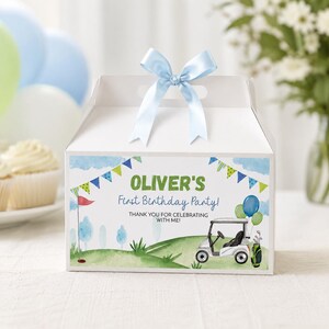 K&ouml;nnte beinhalten: Eine wei&szlig;e Schachtel mit einem hellblauen Satinband oben. Die Vorderseite zeigt ein Golf-Motiv in Aquarell mit gr&uuml;nen H&uuml;geln, einem Golfcart, einer Fahne und Luftballons. Der Text lautet: OLIVER'S First Birthday Party! THANK YOU FOR CELEBRATING WITH ME!