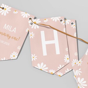 Op de afbeelding: Een decoratieve vlaggenlijn met lichtroze vlaggetjes versierd met witte madeliefjespatronen. De vlaggetjes tonen de tekst 'MILA is turning one!' in wit en goud, de datum '10.08.202X' en een grote witte letter 'H'.