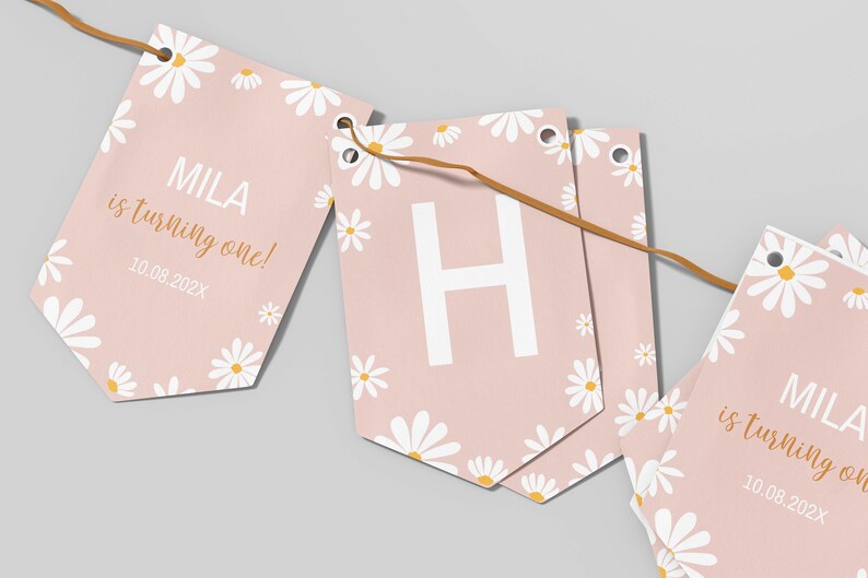 Op de afbeelding: Een decoratieve vlaggenlijn met lichtroze vlaggetjes versierd met witte madeliefjespatronen. De vlaggetjes tonen de tekst 'MILA is turning one!' in wit en goud, de datum '10.08.202X' en een grote witte letter 'H'.