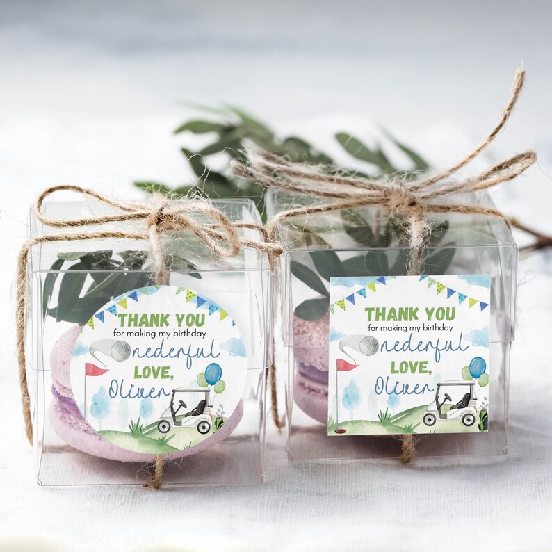 May include: Two clear square plastic favor boxes tied with jute twine, each containing a purple macaron and a green sprig. Attached are circular and square labels featuring a golf cart, balloons, and the text "THANK YOU for making my birthday one-derful LOVE, Oliver."
