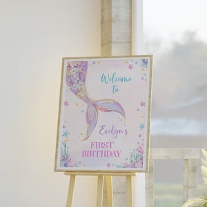 May include: A rectangular sign on a wooden easel featuring a watercolor mermaid tail illustration in purple, gold, and blue. The text reads Welcome to Evelyn's FIRST BIRTHDAY, surrounded by small starfish, coral, and bubbles on a light background.