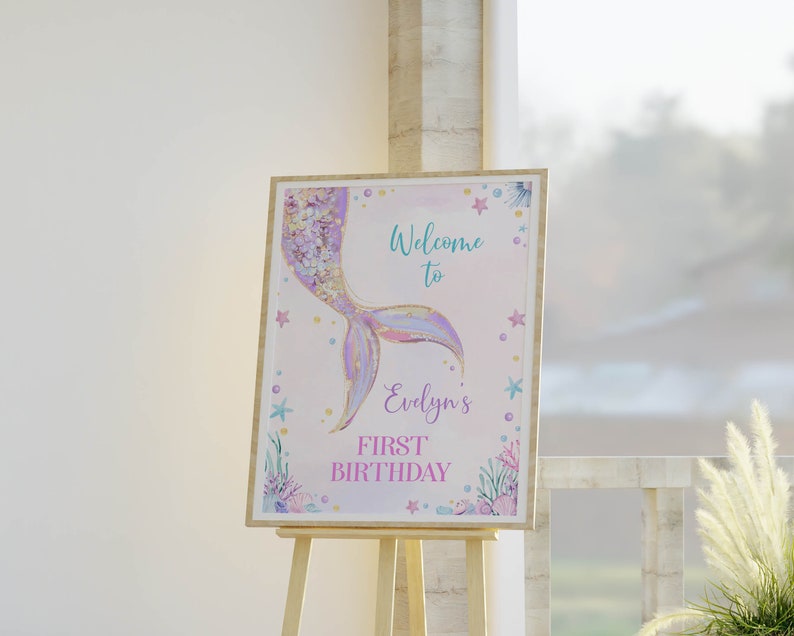 May include: A rectangular sign on a wooden easel featuring a watercolor mermaid tail illustration in purple, gold, and blue. The text reads Welcome to Evelyn's FIRST BIRTHDAY, surrounded by small starfish, coral, and bubbles on a light background.
