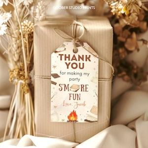 May include: A gift wrapped in brown paper, tied with twine, and adorned with a tag. The tag reads "THANK YOU for making my party S'MORE FUN Love, Jacob" with illustrations of s'mores and a campfire. Dried flowers are in the background.