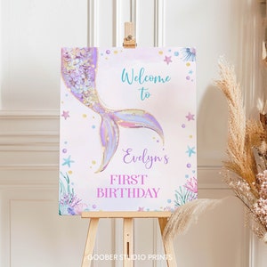 May include: A rectangular sign on a wooden easel featuring a watercolor mermaid tail in shades of purple, blue, and gold. The text reads Welcome to Evelyn's FIRST BIRTHDAY. The design includes small starfish, coral, and bubbles on a light background.