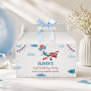 May include: A white cardboard gable box with a light blue satin ribbon bow on top. The front features a watercolor illustration of a red and blue airplane with balloons, blue clouds, and red and blue bunting. Text reads: OLIVER'S First Birthday Party THANK YOU FOR CELEBRATING WITH ME!
