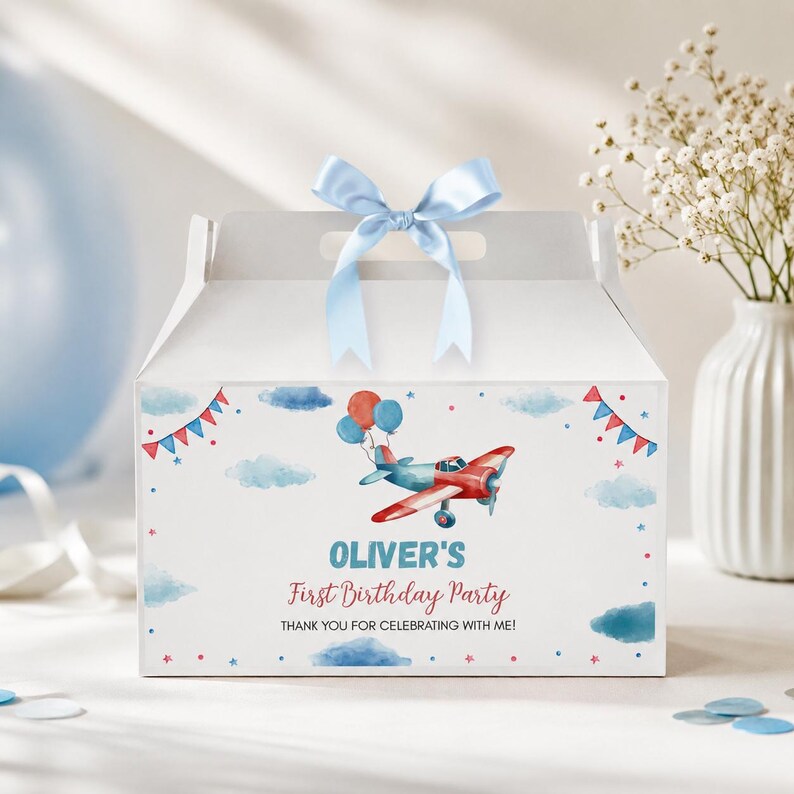 May include: A white cardboard gable box with a light blue satin ribbon bow on top. The front features a watercolor illustration of a red and blue airplane with balloons, blue clouds, and red and blue bunting. Text reads: OLIVER'S First Birthday Party THANK YOU FOR CELEBRATING WITH ME!