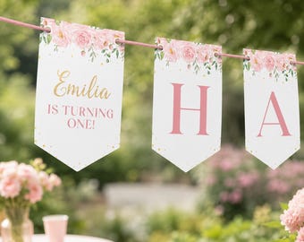 May include: A white pennant banner featuring a pink floral border and gold dots. The first flag displays the text 'Emilia IS TURNING ONE!' in gold and pink script and block lettering. The following two flags show the large pink letters 'H' and 'A'.