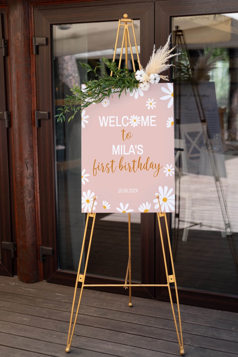 Op de afbeelding: Een rechthoekig roze bord met witte madeliefjesillustraties, met de tekst "WELCOME to MILA'S first birthday" en "10.08.202X". Het bord is versierd met een takje groen en droogbloemen en staat op een gouden metalen driepoot-ezel.