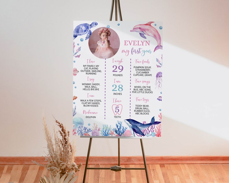 May include: A white milestone birthday sign featuring a circular photo of a child, sea creature illustrations, and text including Evelyn, my first year, 29 pounds, 28 inches, and 5 teeth, surrounded by watercolour coral, a turtle, dolphin, and whale.