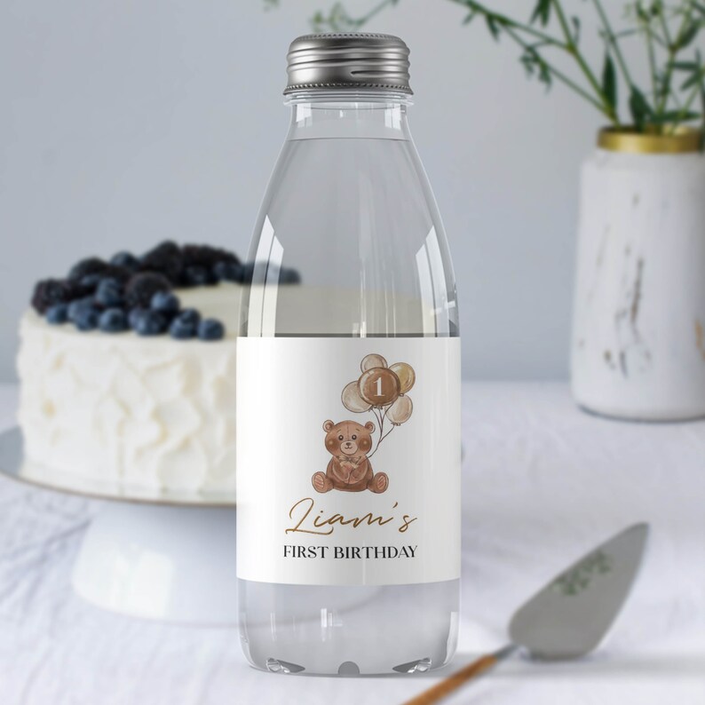 May include: A clear plastic water bottle with a silver screw cap and a white label featuring a brown teddy bear illustration holding a cluster of balloons with the number 1, the text Liam's, and the text FIRST BIRTHDAY in black and gold.