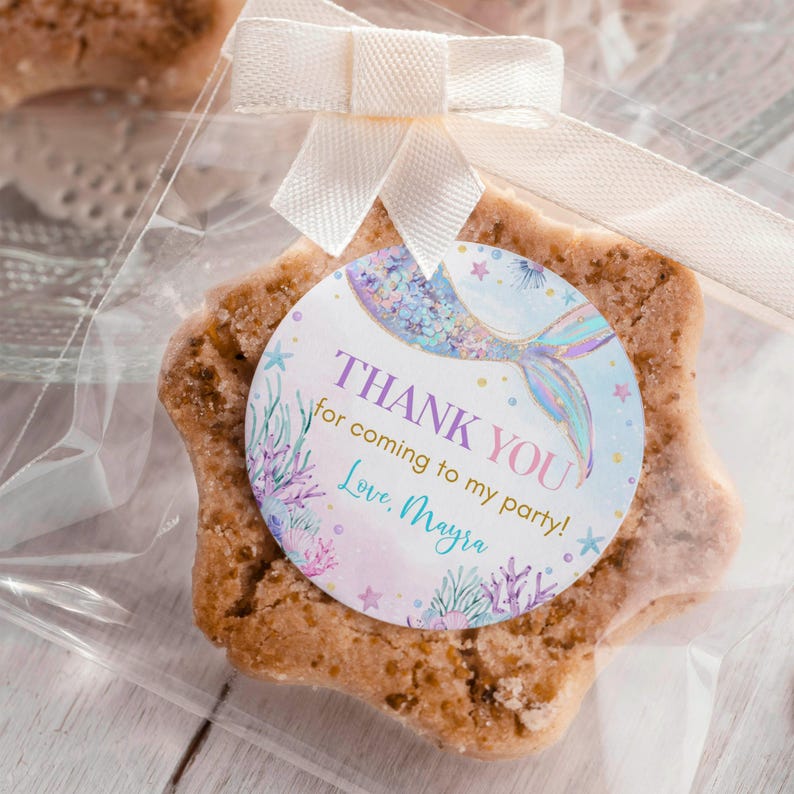 May include: A star-shaped cookie with a round sticker featuring a mermaid tail with glitter and a thank you message. The sticker says "Thank You! for coming to my party! Love, Mayra".