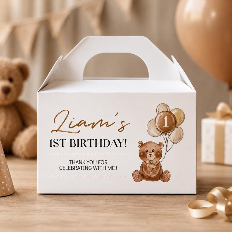 May include: A white cardboard gable box featuring a brown teddy bear illustration holding a cluster of balloons with the number 1. The text reads Liam's 1ST BIRTHDAY! THANK YOU FOR CELEBRATING WITH ME! The box has a top handle and a clean, rectangular shape.