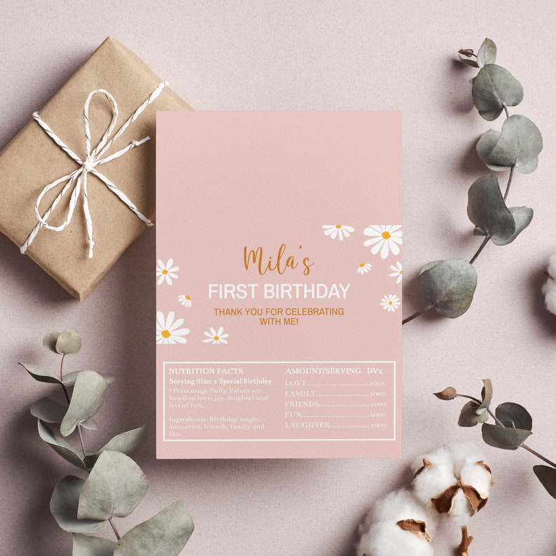 Op de afbeelding: Een roze printbare kaart met witte madeliefjes en gouden tekst die "Mila's First Birthday" en "Bedankt voor het vieren met me!" zegt. De kaart bevat ook een sectie met voedingsinformatie met de tekst "Serving Size: a Special Birthday" en "Amount/Serving DV%" met een lijst van items zoals "Love", "Family", "Friends", "Fun" en "Laughter".
