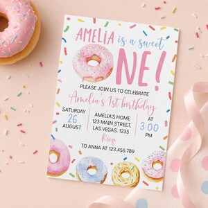 May include: A rectangular birthday invitation card featuring watercolor donut illustrations and colorful sprinkles. The text reads: AMELIA is a sweet ONE! PLEASE JOIN US TO CELEBRATE Amelia's 1st birthday SATURDAY 26 AUGUST AMELIA'S HOME 123 MAIN STREET, LAS VEGAS. 1235 AT 3:00 PM RSVP TO ANNA AT 123.456.789.