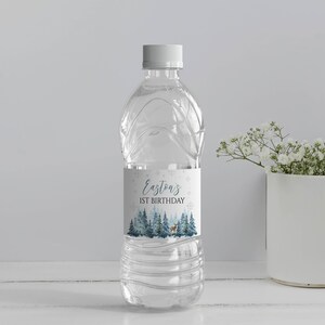 May include: A clear plastic water bottle with a white cap and a custom label featuring a winter forest scene with pine trees, a small deer, and snowflakes. The text on the label reads Easton's 1ST BIRTHDAY in dark blue and black lettering.