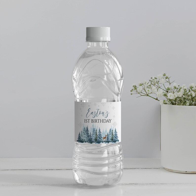 May include: A clear plastic water bottle with a white cap and a custom label featuring a winter forest scene with pine trees, a small deer, and snowflakes. The text on the label reads Easton's 1ST BIRTHDAY in dark blue and black lettering.