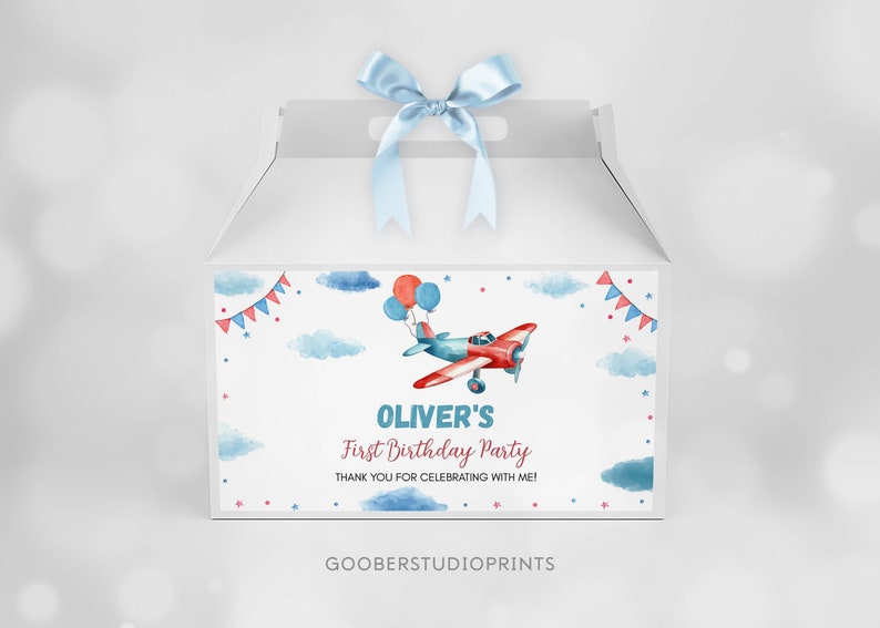 May include: A white gable box with a light blue satin ribbon bow on top. The front features a watercolor illustration of a red and blue airplane with balloons, clouds, and bunting. Text reads: OLIVER'S First Birthday Party THANK YOU FOR CELEBRATING WITH ME!