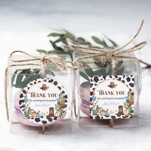 May include: Two clear square favor boxes tied with jute twine, each containing a light purple macaron and greenery. Each box features a western-themed sticker with a cow print border, a cowboy hat, cactus, and the text Thank you for coming to my party! Love, Oliver.