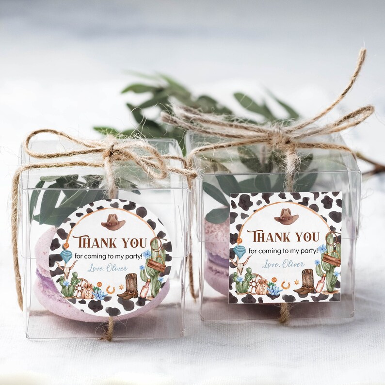 May include: Two clear square favor boxes tied with jute twine, each containing a light purple macaron and greenery. Each box features a western-themed sticker with a cow print border, a cowboy hat, cactus, and the text Thank you for coming to my party! Love, Oliver.