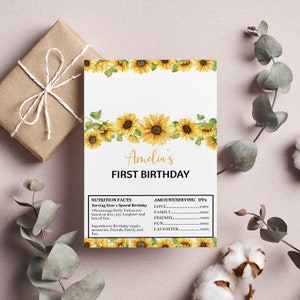 May include: A rectangular birthday card featuring a sunflower border at the top and bottom. The text reads Amelia's FIRST BIRTHDAY. Below is a nutrition facts label listing ingredients like birthday magic and values for love, family, friends, fun, and laughter.