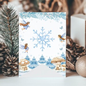 May include: A rectangular card featuring a central light blue snowflake with a heart-shaped center, surrounded by illustrations of winged fairies, blue pine trees, mushrooms, and butterflies against a white background with pine branch borders.