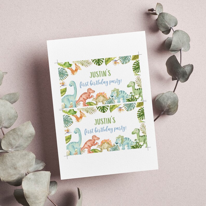 May include: A rectangular white card featuring two identical prints of a dinosaur-themed birthday invitation. The text reads Justin's first birthday party! surrounded by watercolor illustrations of various dinosaurs and tropical green leaves.