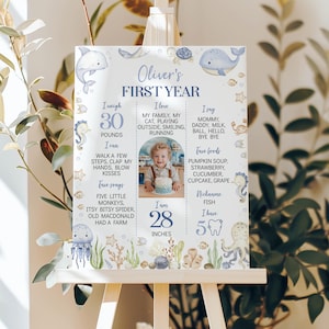 May include: A white milestone board titled Oliver's First Year featuring blue text, a central photo of a child with a cake, and watercolor illustrations of sea creatures including a dolphin, whale, jellyfish, octopus, and seahorses.