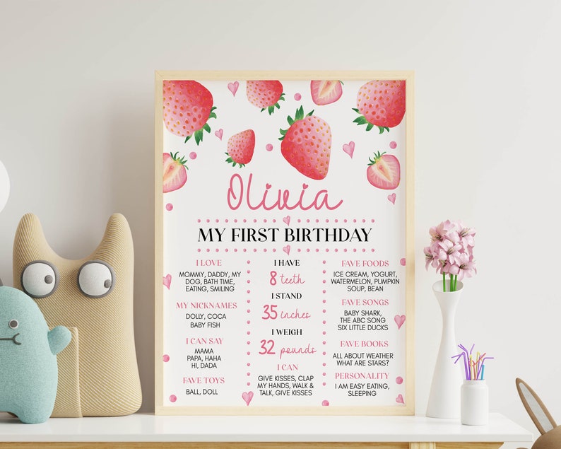 May include: A rectangular birthday milestone poster in a light wood frame featuring strawberry illustrations, pink hearts, and dots. The text includes the name Olivia, My First Birthday, and lists personal details like 8 teeth, 35 inches, and 32 pounds.