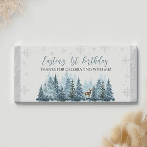 May include: A rectangular candy bar wrapper featuring a watercolor forest of blue and green pine trees with a small brown deer. The text reads Easton's 1st birthday THANKS FOR CELEBRATING WITH ME! surrounded by light blue snowflake illustrations.