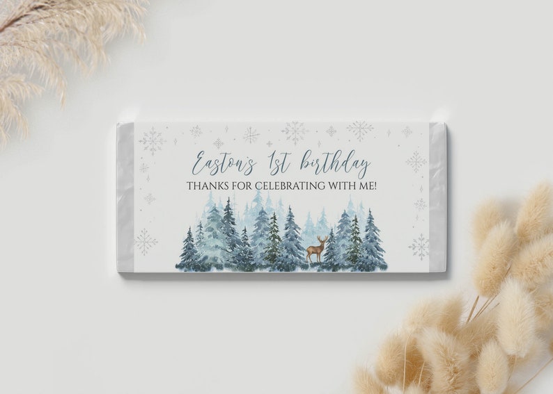 May include: A rectangular candy bar wrapper featuring a watercolor forest of blue and green pine trees with a small brown deer. The text reads Easton's 1st birthday THANKS FOR CELEBRATING WITH ME! surrounded by light blue snowflake illustrations.