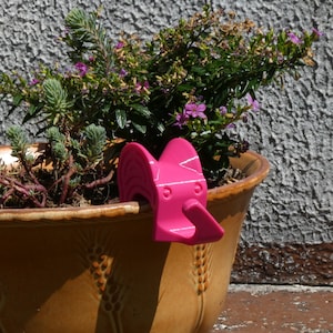 Könnte beinhalten: Ein rosafarbener, elefantenförmiger Pflanzenhalter aus Plastik ist am Rand eines Terrakotta-Blumentopfs befestigt. Der Topf ist mit grünen Pflanzen und Blumen gefüllt.