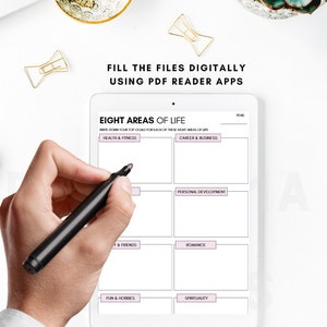 May include: A white tablet with a grid of eight boxes, each labeled with a different area of life: Health & Fitness, Career & Business, Personal Development, Y & Friends, Romance, Fun & Hobbies, Spirituality, and Year. A hand is holding a black marker pen over the tablet.