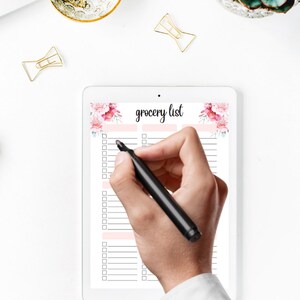May include: A white tablet showing a "grocery list" with floral decorations. A hand with a black marker is about to write on the list. Gold paper clips and a small decorative bowl are on the white surface.