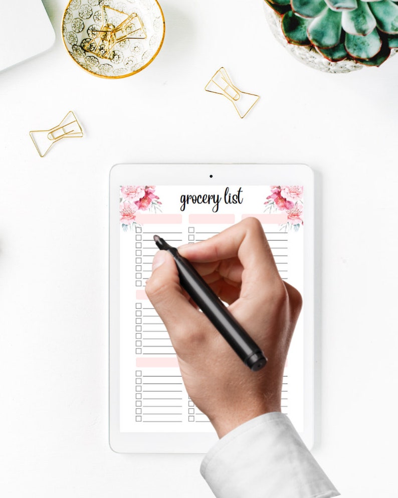 May include: A white tablet showing a "grocery list" with floral decorations. A hand with a black marker is about to write on the list. Gold paper clips and a small decorative bowl are on the white surface.
