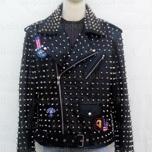 Puede incluir: Chaqueta de cuero negra con tachuelas cónicas plateadas, cuello con solapas y cintura con cinturón. La chaqueta presenta cierre de cremallera, múltiples bolsillos y parches con diseños gráficos.