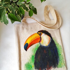 May include: A white canvas tote bag featuring a hand-painted toucan design. The toucan has a large orange and yellow beak and black feathers. The background is green with white splatters of paint. The bag has two long straps and the artist's signature "a.s.bog" is in the bottom right corner.
