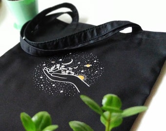 May include: Black tote bag with a white design of a hand holding a small house, a moon, stars, and planets. The design is in the center of the bag.
