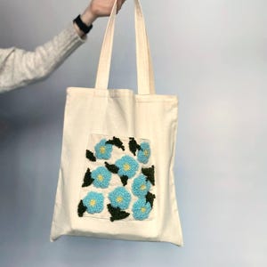 Op de afbeelding: Een witte canvas tote bag met een blauw en groen bloemenpatroon geborduurd op de voorkant. Het ontwerp bevat een cluster van blauwe bloemen met groene bladeren.