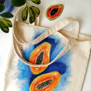 May include: A white canvas tote bag with a blue background and two painted papaya halves. The papaya halves are orange and yellow with black seeds. The bag has two white straps.
