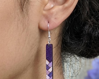 May include: A drop earring featuring a long, rectangular pendant with a dark purple glitter top section and a bottom section displaying a geometric, woven-style pattern in alternating shades of light purple, lavender, and white, suspended by a silver hook.
