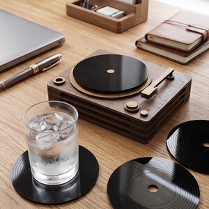 May include: A set of black circular drink coasters designed to resemble miniature vinyl records, with one coaster supporting a clear glass filled with ice water and two additional coasters placed nearby on a wooden desk surface.
