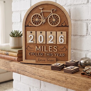May include: An arched wooden tracker sign featuring a laser-cut bicycle icon at the top, with four slots displaying the number 2026. Below, the text reads MILES CYCLED THIS YEAR, with a nameplate at the bottom engraved with the name Karen Turner.