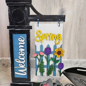 May include: Black wooden welcome sign with a blue background and the word "Welcome" written in white. The sign is attached to a black metal post with a white sign that says "Spring" in yellow letters. The sign is decorated with a white picket fence, flowers, and a watering can.