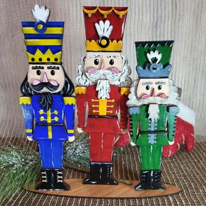 May include: Three wooden nutcracker figurines standing on a wooden base. The nutcracker on the left is wearing a blue and yellow uniform with a black beard. The nutcracker in the middle is wearing a red and yellow uniform with a white beard. The nutcracker on the right is wearing a green and gray uniform with a white beard.