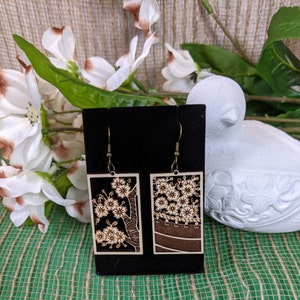 May include: A pair of wooden earrings with a cherry blossom design. The earrings are rectangular and have a brown background with white cherry blossoms. The earrings are hanging from a black display card.